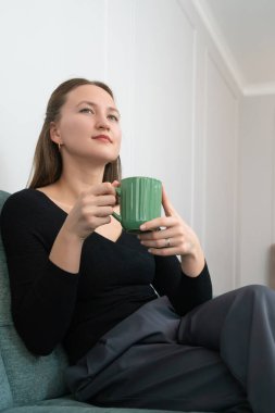 Siyah bir kazak giyen genç bir kadın rahat bir şekilde kanepede oturuyor, rahat ve davetkar bir atmosferde şık yeşil bir bardaktan sıcak bir içecek içiyor. Dikey fotoğraf