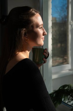Yeşil bir kupa tutan genç bir kadın, parlak, güneşli bir günde pencereden dışarı bakıyor, huzur ve huzur dolu bir anın tadını çıkarıyor. Dikey fotoğraf
