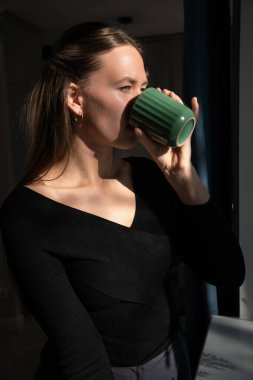 Siyah bluzlu genç bir kadın pencerenin kenarındaki yeşil bardaktan yudumlarken huzurlu bir anın tadını çıkarıyor. Güneş ışığı yavaşça yüzünü aydınlatıyor ve sakin bir atmosfer yaratıyor.