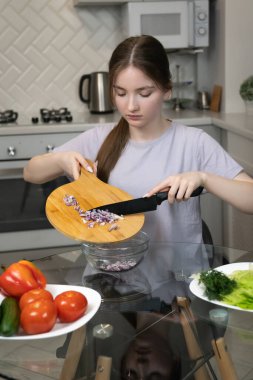 Genç bir kız, domates, salatalık ve marul gibi taze sebzelerle çevrili modern mutfakta dilimlenmiş kırmızı soğanı kaseye doldurarak sağlıklı yemek ve yaşam tarzını ön plana çıkarıyor.