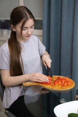 İşine odaklanan genç bir kadın, modern mutfağında besleyici bir yemek için taze kırmızı dolma biberleri ahşap bir tahtada ustalıkla kesiyor. Dikey fotoğraf