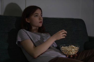 Young girl sitting on a sofa is watching a scary movie on television in a dark room, eating popcorn from a glass bowl and covering her mouth with her hand