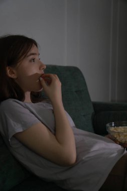 Teenage girl is lying on a comfortable sofa in her living room at night, eating popcorn from a glass bowl and watching a movie on television. Vertical photo