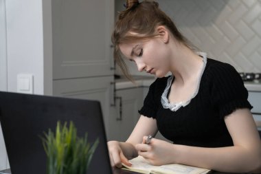Young woman using laptop and taking notes in notebook, studying online in modern kitchen, creating comfortable and productive learning environment at home