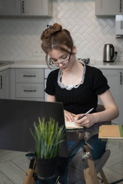 Young woman taking notes while studying on her laptop, writing in a notebook, and engaging in online learning from the comfort of her cozy kitchen, showcasing focus and concentration