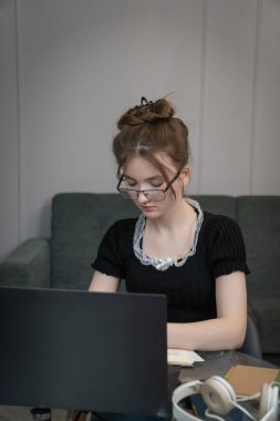Young student girl wearing glasses is taking notes while attending online class or e-learning course, using laptop at home, focused on her studies and education. Vertical photo