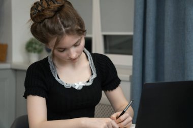 Focused young female student is taking notes from her laptop, preparing for exams or doing homework, demonstrating the concept of online education and self-discipline