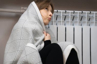 Young woman shivering under a blanket beside a white radiator, worried about high heating bills and energy shortage during a chilly winter evening at home