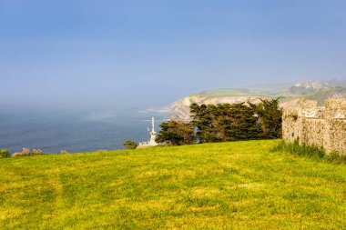 Kırsal Cantabria manzarası: Sarı çiçekler ve taş duvarlarla kaplı çimenler, sisli okyanuslar, dağlar ve arka planda küçük bir kasaba. Comillas, Cantabria, İspanya.