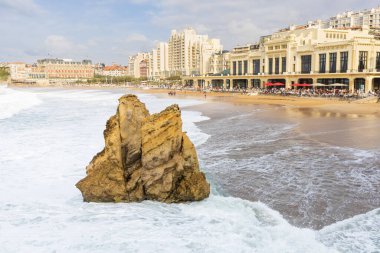 Köpüren dalgalar La Grande Plage kumsalına çarpıyor. Sarı bir kaya ve arka planda şehrin binaları var. Biarritz, Pireneler-Atlantikler, Fransa.