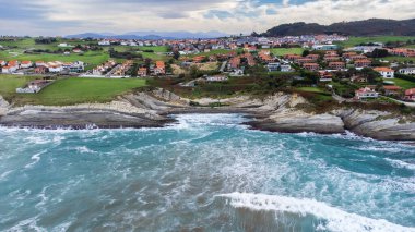 Kıyı şeridinin alışılmadık katmanlı kaya oluşumları, fırtınalı Atlantik Okyanusu, küçük bir köy ve sisin içindeki dağlar. Costa Quebrada Geopark, Cantabria, İspanya.