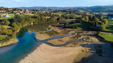 Ria de la Rabia nehrinin kumlu adalarıyla çevrili, çimen ve ağaçlarla kaplı yeşil tepeler ve tepenin üzerine tünemiş küçük bir köy. Oyambre Doğal Parkı, La Rabia, Comillas, Cantabria, İspanya.