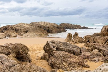 Alışılmadık kaya oluşumları ve kayalık taşlar bulutlu bir gökyüzünün altındaki kumlu Playa Liencres plajını (Playa de Valdearnas) işaret eder. Liencres, Cantabria, İspanya.