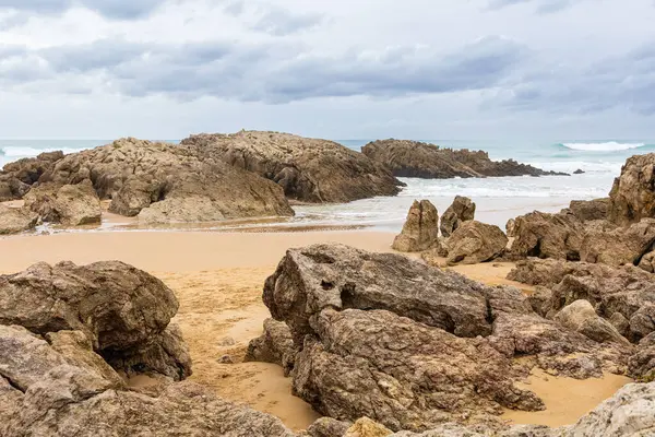 Alışılmadık kaya oluşumları ve kayalık taşlar bulutlu bir gökyüzünün altındaki kumlu Playa Liencres plajını (Playa de Valdearnas) işaret eder. Liencres, Cantabria, İspanya.