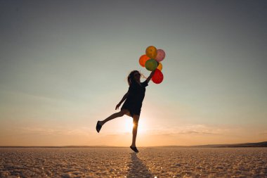 Gün batımında kumsalda gökyüzüne karşı duran balonları tutan kadın silueti.