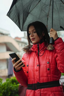 Genç ve güzel bir kadın şemsiye tutarken ve yağmurlu bir günde şehirde yürürken akıllı telefon kullanıyor.