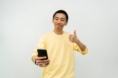 Akıllı telefonlu genç Asyalı adam beyaz stüdyoda baş parmak gösteriyor. 