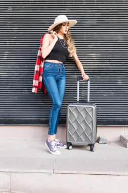 Hasır şapka ve damalı tişörtle bavulla poz veren güzel kız.