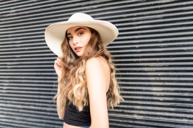 Genç bir kadın şehirde kameranın önünde hasır şapkayla poz veriyor.