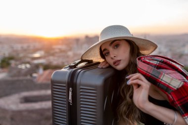 Hasır şapkalı kadın, günbatımının arka planında bavul taşıyor.