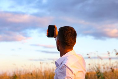 Günbatımında gökyüzü geçmişi olan bir adam telefonla fotoğraf çekiyor.