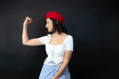 Kırmızı şapkalı ve beyaz gömlekli bir kadın kolunu esnetiyor. Görüntünün güçlü, kendinden emin bir havası var.
