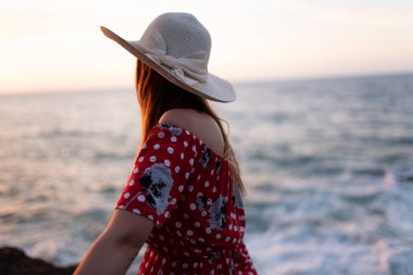 Hasır şapkalı bir kadın okyanusun yakınındaki bir kayanın üzerinde oturuyor..