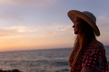 Hasır şapkalı bir kadın okyanusun yakınındaki bir kayanın üzerinde oturuyor..