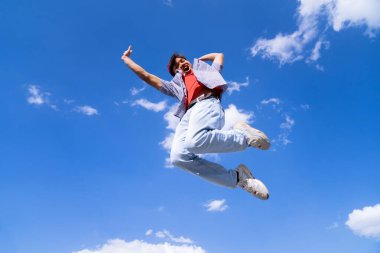 Kolları açık bir adam havaya zıplıyor. Gökyüzü mavi ve arka planda bulutlar var. Manzara neşeli ve enerjik.