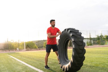 Bir adam tarlada lastik kaldırıyor. Kolye takıyor ve kaslı bir yapısı var.