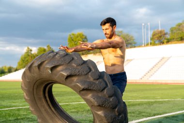 Bir adam tarlada lastik kaldırıyor. Kolye takıyor ve kaslı bir yapısı var.