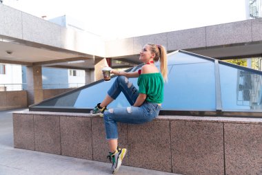 Genç bir kadın şehirde kağıt bardakla kahve içiyor.