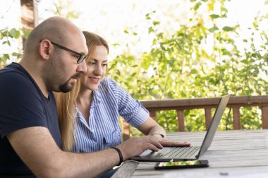 Genç ve etnik çeşitlilikte mutlu bir çift parkta birlikte vakit geçiriyorlar, masada dizüstü bilgisayarla otururken ders çalışıyorlar.