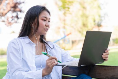 Genç ve güzel Asyalı bir kadın internette / laptopta çalışıyor açık hava parkında alışveriş yapıyor.,