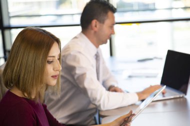 Bir kadın ve bir adam laptopları ve tabletleriyle bir masada oturuyorlar.
