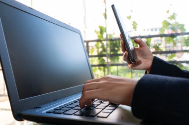 Bir insan elinde cep telefonu tutarken dizüstü bilgisayarda yazı yazıyor.