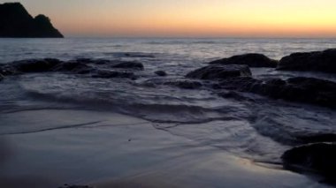 Parlak deniz günbatımı. Dalgalar kayaya çarpıyor, gün batımının sıcaklığıyla, kum ve çakıl taşlarıyla., 