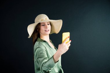 Hasır şapkalı bir kadın cep telefonuyla selfie çekiyor. Gülümsüyor ve eğleniyor.