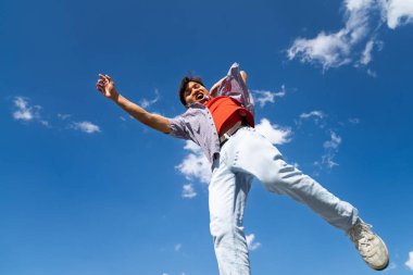 Kolları açık bir adam havaya zıplıyor. Gökyüzü mavi ve arka planda bulutlar var. Manzara neşeli ve enerjik.