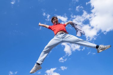 Kolları açık bir adam havaya zıplıyor. Gökyüzü mavi ve arka planda bulutlar var. Manzara neşeli ve enerjik.