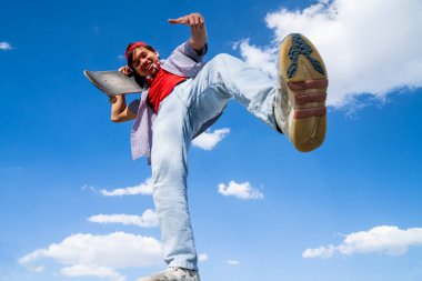 Genç adam arka planda mavi ve bulutlu bir kaykayla mutlu poz veriyor. gençlik sporları ve eğlence konsepti