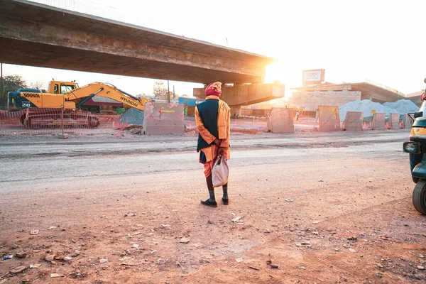 Old Indian man wearing a red turban and orange dhoti kurta with jacket standing and waiting for transport vehicle on main road, An old man standing on beside road construction, beautiful sunlight