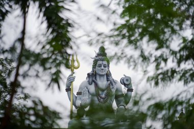 Büyük Lord Shiva heykeli yeşil ağaç dallarının arasından görülüyor, Hindu tanrısı Shiva trishul ile çevrili, doğal yeşilliklerle çevrili, Ruhsal Lord Shiva heykeli açık hava manzaralı, görkemli Shiva putu, kısmen ağaçlar tarafından gizlenmiş, mızrak tutan..