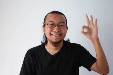 Yetişkin Asyalı adam siyah tişört ve uzun saçlı gözlük takıyor. Gri arka planda el işareti yapıyor..