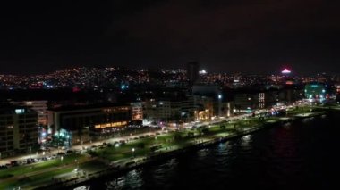 İzmir şehrinin gece manzarası, deniz kenarındaki binalar ve sahildeki insanlar. İzmir 'deki gezinti güvertesi boyunca İHA atışı. Yüksek kalite 4k görüntü