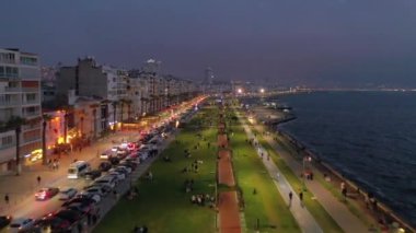 İzmir şehrinin gece manzarası, deniz kenarındaki binalar ve sahildeki insanlar. İzmir 'deki gezinti güvertesi boyunca İHA atışı. Yüksek kalite 4k görüntü