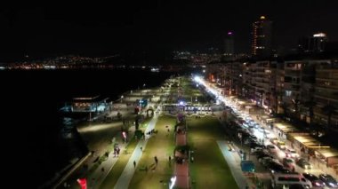 İzmir şehrinin gece manzarası, deniz kenarındaki binalar ve sahildeki insanlar. İzmir 'deki gezinti güvertesi boyunca İHA atışı. Yüksek kalite 4k görüntü