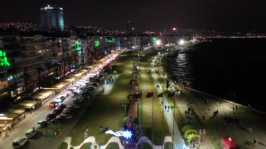 İzmir şehrinin gece manzarası, deniz kenarındaki binalar ve sahildeki insanlar. İzmir 'deki gezinti güvertesi boyunca İHA atışı. Yüksek kalite 4k görüntü