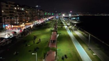 İzmir şehrinin gece manzarası, deniz kenarındaki binalar ve sahildeki insanlar. İzmir 'deki gezinti güvertesi boyunca İHA atışı. Yüksek kalite 4k görüntü
