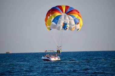 2 people Parasailing in blue sky, towed by a boat. Multicloured parasail. Coming into land by the boat.  clipart